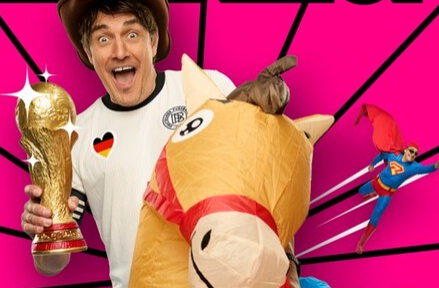 Ein fröhlicher Mann hält den Fußball-Weltpokal und steht neben einem aufblasbaren Pferd. Der Hintergrund ist bunt und dynamisch gestaltet.