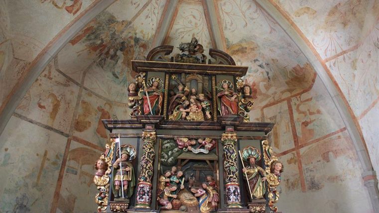 Ein kunstvoll geschnitzter Altar in einer Kirche, reich verziert mit Figuren und religiösen Darstellungen. Die Wände sind mit alten Wandmalereien geschmückt.