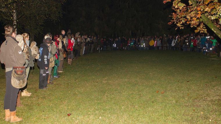 Een grote groep mensen staat in de open lucht, omringd door bomen, 's nachts. De aanwezigen lijken te wachten op een evenement of bijeenkomst.