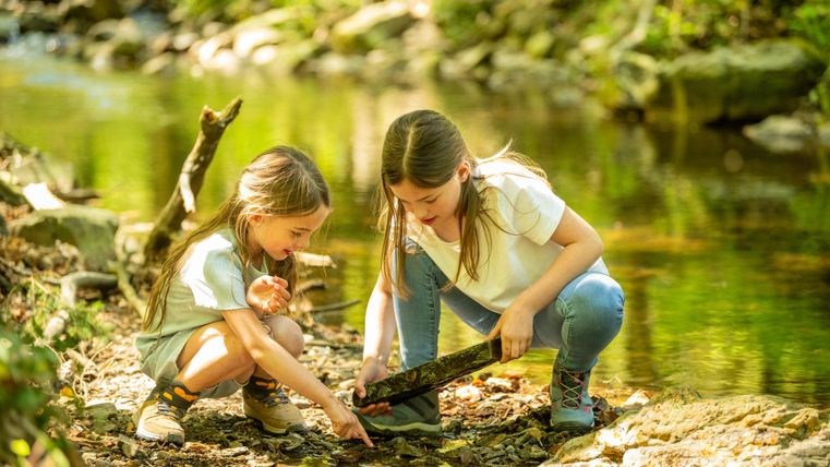 Two girls are playing by the bank of a small stream. They are examining a stick in the water together.