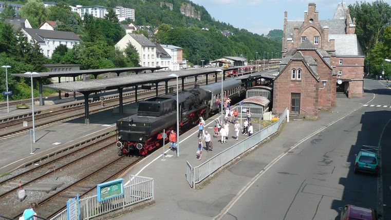 Gerolstein station with a steam locomotive on the tracks. Travelers are walking across the platform. Historical station building in the background.