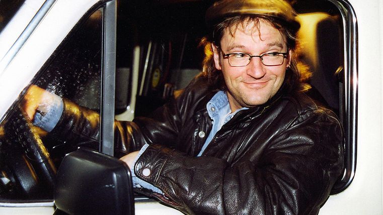 A man sits smiling behind the wheel of a vehicle. He is wearing a leather jacket and a cap.