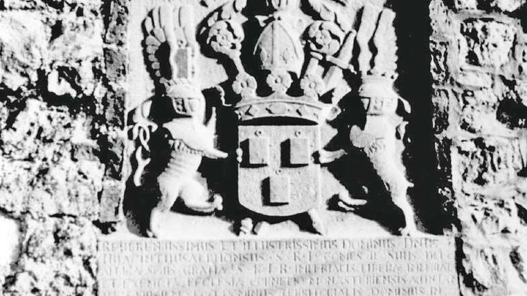 A coat of arms with a royal shield and two lions holding it. The background consists of a stone wall.