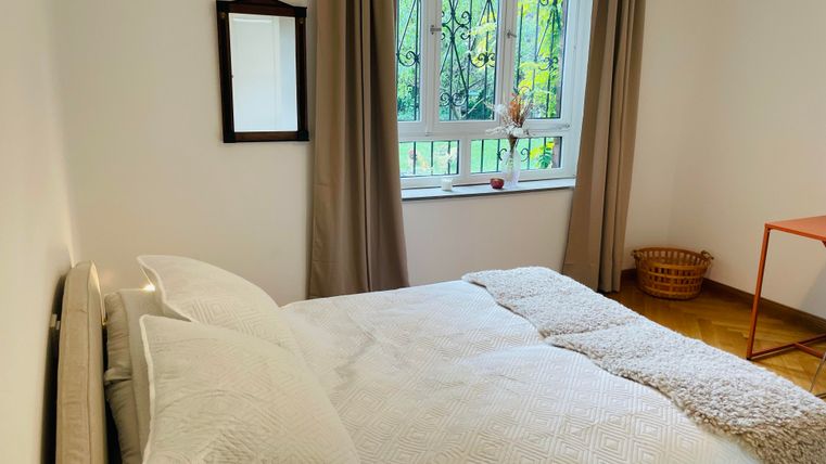 Une chambre confortable avec un grand lit et des rideaux clairs. Une fenêtre laisse entrer beaucoup de lumière du jour et une petite table se trouve à côté.