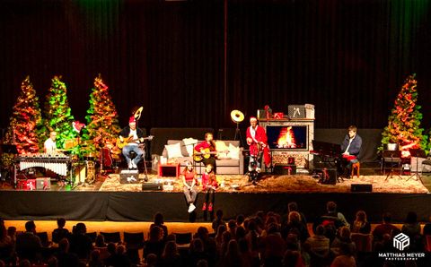 Een muzikaal kerstconcert op het podium met meerdere muzikanten en kerstversiering. Op de achtergrond zijn feestelijk versierde dennenbomen te zien.