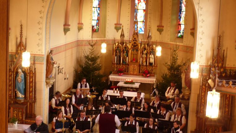 Ein Chor singt in einer festlich dekorierten Kirche. Im Hintergrund sind Weihnachtsbäume und farbenfrohe Fenster zu sehen.