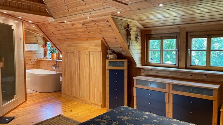 A cozy bedroom with a wooden ceiling and large windows. In the background, a modern bathroom with a bathtub can be seen.