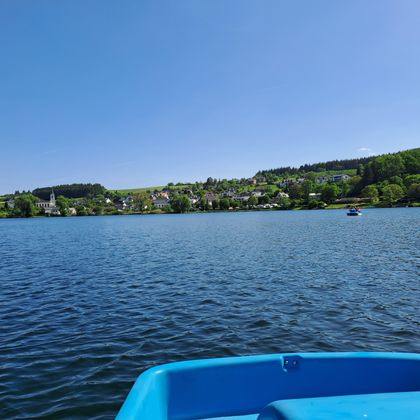 A tranquil lake with clear water and a beautiful shoreline. In the background, green hills and some buildings are visible.