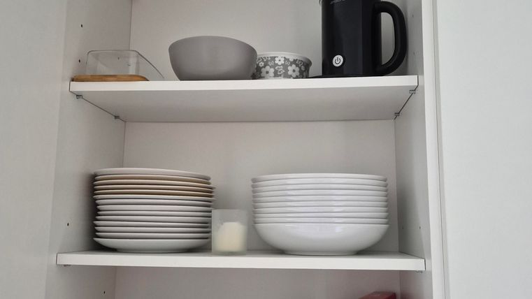 Een open keukenkast met verschillende serviesdelen. Boven staan kopjes en een koffiezetapparaat, terwijl beneden borden en kommen ordelijk gesorteerd zijn.