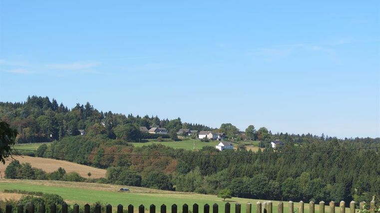 Eine ruhige Landschaft mit sanften Hügeln und verstreuten Häusern. Der Himmel ist klar und blau, umgeben von grünen Wäldern und Feldern.