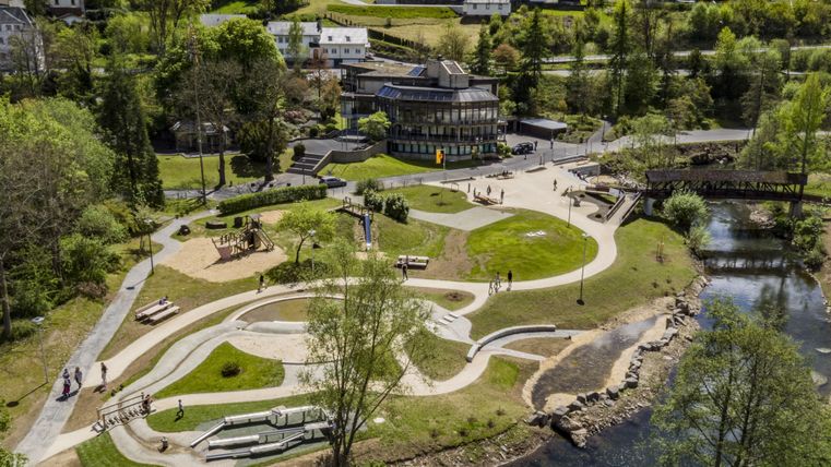 Eine grüne Parklandschaft mit Wegen, Bäumen und einem Fluss. Im Hintergrund sind Gebäude und eine Aussichtsplattform zu sehen.