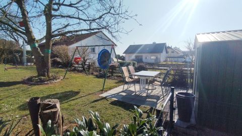 Un jardin paisible avec une balançoire et un coin salon confortable. En arrière-plan, plusieurs maisons et un ciel bleu clair sont visibles.