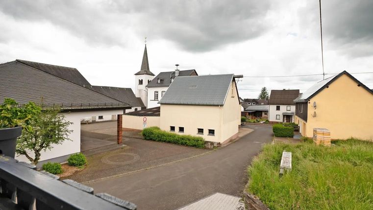 Ein ruhiges Dorf mit mehreren Häusern und einer Kirche im Hintergrund. Der Himmel ist bewölkt und es gibt viel Grün in der Umgebung.