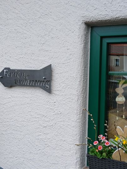Ein Schild mit der Aufschrift "Ferienwohnung" zeigt den Eingang. Neben dem Fenster stehen Blumen in einem Topf.