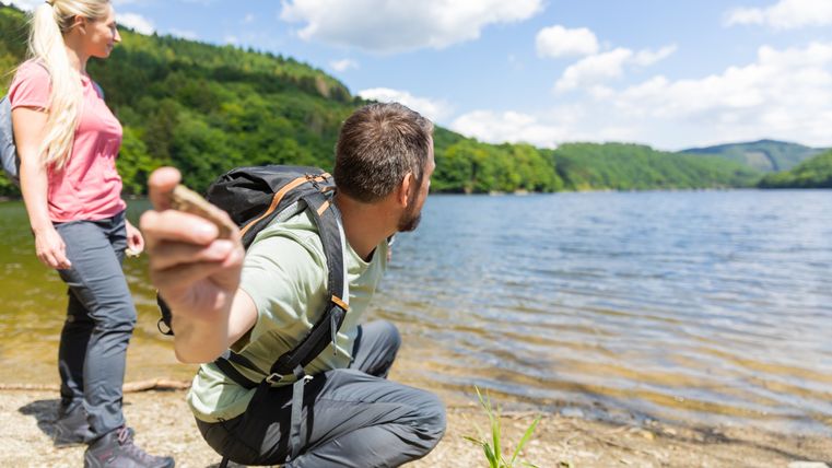 Zwei Personen am Ufer eines Sees, eine kniet und hält einen Stein, die andere steht. Im Hintergrund sind Bäume und blauer Himmel zu sehen.