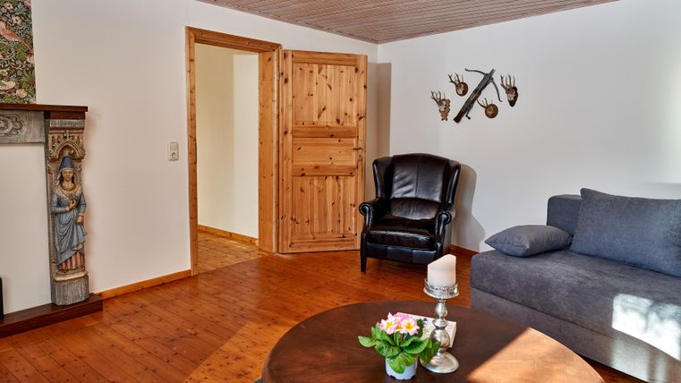 Ein gemütliches Wohnzimmer mit einem schwarzen Sessel, einem grauen Sofa und einem runden Tisch. Die warme Holzverkleidung und die dekorativen Elemente verleihen dem Raum einen einladenden Charakter.