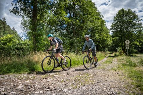 Twee fietsers rijden op een grindpad in een groen bosgebied. De lucht is gedeeltelijk bewolkt en de omgeving oogt uitnodigend en natuurlijk.