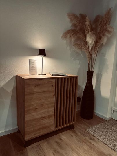 Une armoire en bois élégante se trouve à côté d'une lampe décorative. En arrière-plan, il y a un grand vase avec des fleurs sèches.