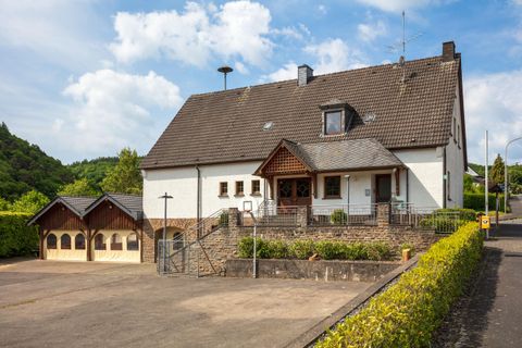 Een groot, licht huis met een garageaanbouw en een verzorgde voortuin. De omgeving is groen en rustig, met een blauwe lucht op de achtergrond.