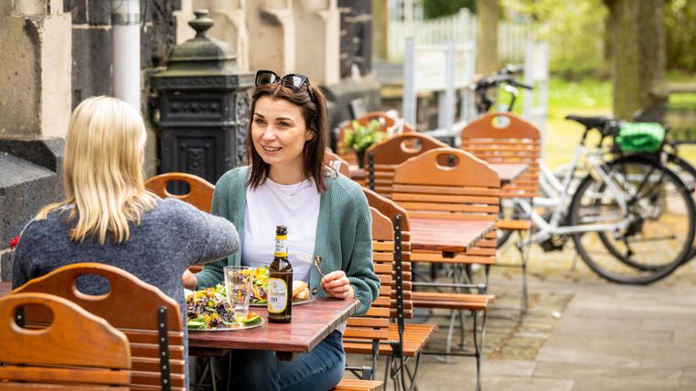 Zwei Frauen sitzen an einem Tisch im Freien und genießen Getränke und Snacks. Im Hintergrund sind Fahrräder und eine grüne Umgebung zu sehen.