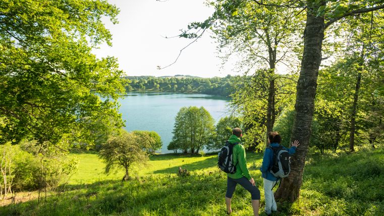 Two hikers look out over the Weinfelder Maar, a lake surrounded by meadows and trees.