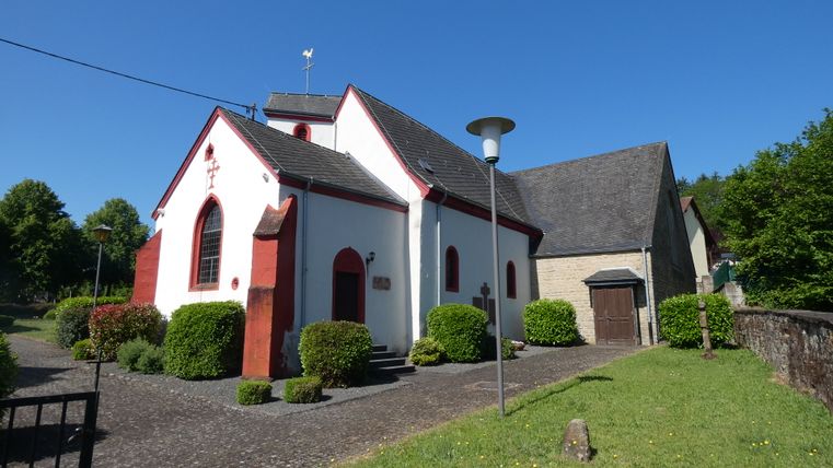 White church with red accents and a gray roof in Dahlem, surrounded by bushes and trees, in sunny weather.