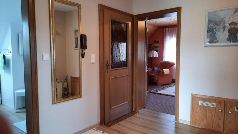 Une entrée lumineuse avec un miroir et une porte intérieure en bois. À l'arrière-plan, un salon confortable avec un canapé rouge est visible.