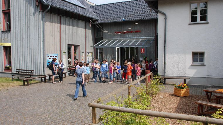 Eine Gruppe von Menschen steht vor einem modernen Gebäude mit Holzwänden. Im Vordergrund sind Bänke und Blumenbeete sichtbar.