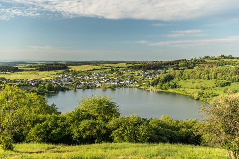 Ein malerischer Blick auf das Schalkenmehrener Maar in der Vulkaneifel bei sonnigem Wetter. Im Vordergrund dichter, grüner Laubwald, dahinter der kreisrunde Maarsee mit spiegelnder Wasseroberfläche. Umgeben ist der See von Wiesen, Feldern und dem kleinen Ort Schalkenmehren. Im Hintergrund erstreckt sich die hügelige Eifellandschaft unter einem blauen Himmel mit leichten Schleierwolken.