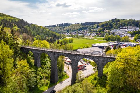 Un impressionnant pont en pierre s'élève au-dessus d'un paysage verdoyant. Entouré de forêts et de petits bâtiments, il donne à l'image une atmosphère charmante.