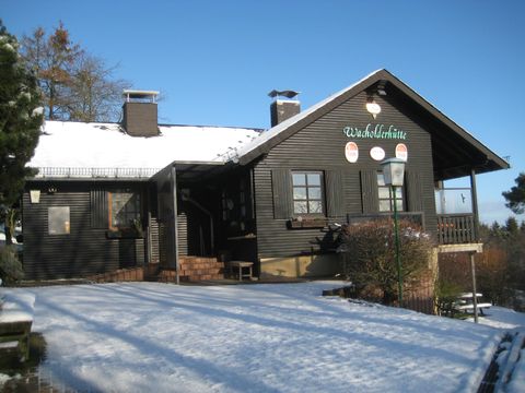 Een zwart houten huis met witte ramen ligt in de sneeuw. De lucht is helder en blauw.