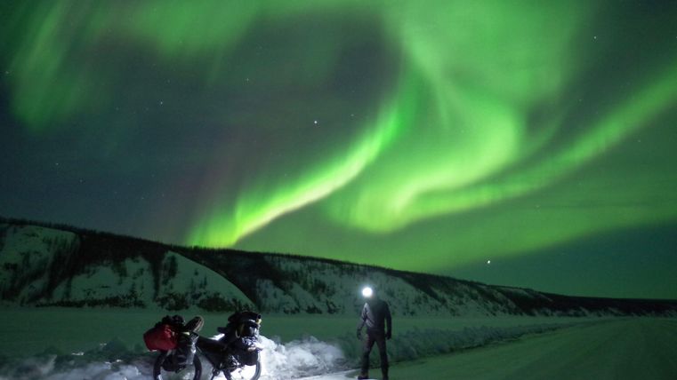 An impressive Northern Lights hover over a snowy landscape. Two people with bicycles stand beneath the luminous sky.