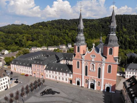 An impressive church with two towers and a beautiful red facade. In the background, green hills and a bustling cityscape can be seen.