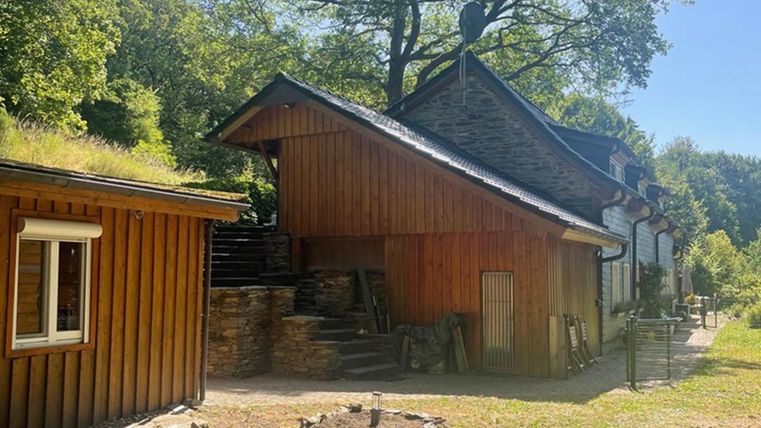 A rustic wooden house with a covered roof next to another building. The garden is green and surrounded by trees.