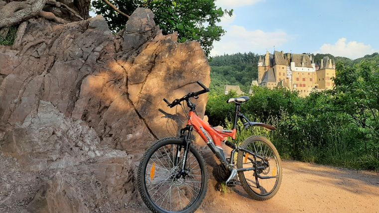 A mountain bike stands next to a large rock on a path. In the background, a castle and a green landscape can be seen.