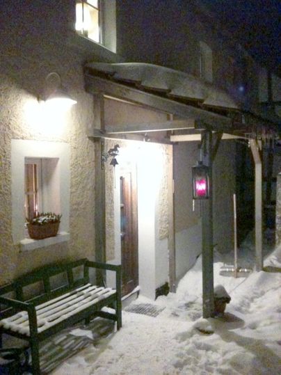 Een gezellige ingang met een bank en een verlichte lantaarn. De grond is bedekt met sneeuw en er heerst een nachtelijke sfeer.