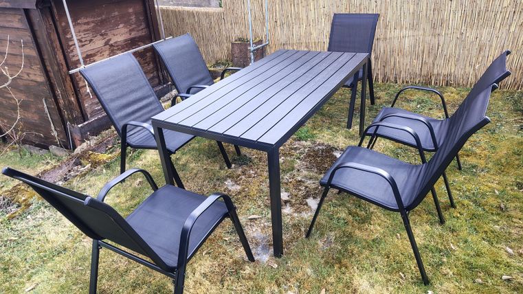 A garden table with six black chairs stands on green grass. In the background, a wooden wall is visible.