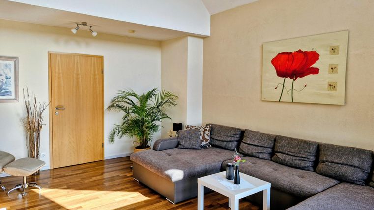 Een moderne woonkamer met een grote grijze bank en een witte tafel. Aan de wand hangt een schilderij van een rode bloem, en er staat een plant in de hoek.