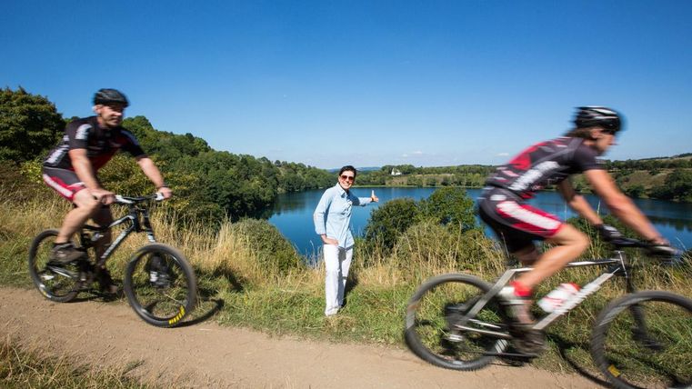 Zwei Mountainbiker fahren entlang eines Pfades mit Blick auf einen ruhigen See. Im Vordergrund steht eine Person, die freundlich auf den See zeigt.