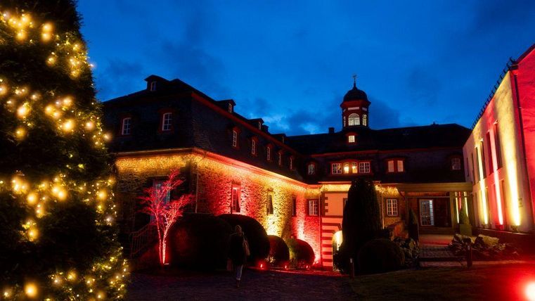Ein festlich beleuchtetes Gebäude bei Nacht, umgeben von schönen Pflanzen. Ein großer Weihnachtsbaum mit Lichtern steht im Vordergrund.