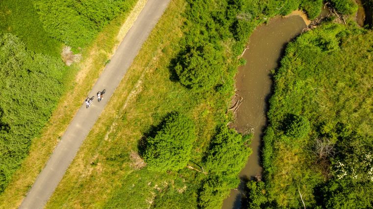 Eine grüne Landschaft mit einem schmalen Weg und einem kleinen Fluss. Einige Personen sind entlang des Weges unterwegs.