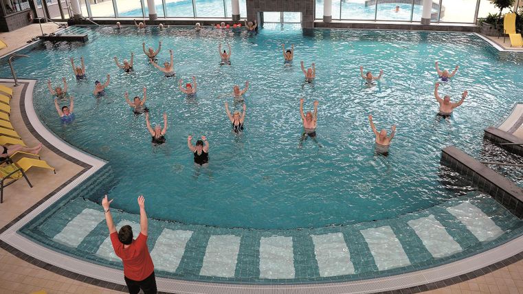 Eine Gruppe von Menschen macht Wassergymnastik in einem hellen Schwimmbad. Im Hintergrund sind große Fenster und ein angenehmes Ambiente zu sehen.