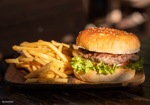 Ein saftiger Burger mit Sesambrötchen, Salat und Tomate liegt auf einem Holzbrett. Daneben befinden sich knusprige Pommes frites.