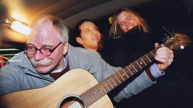 Three men sit in a car while one plays the guitar. All three are having fun and laughing.