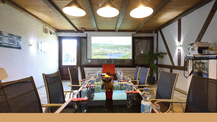 A modern conference room with a long table and chairs. On the wall, there is a projector, and the window offers a view outside.