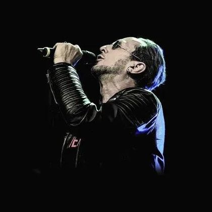 Ein Sänger tritt auf, während er ein Mikrofon hält. Er trägt eine schwarze Lederjacke und Sonnenbrillen.