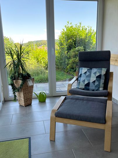Ein bequemer Sessel steht vor großen Fenstern mit Blick auf einen grünen Garten. Neben dem Sessel befindet sich ein Topf mit einer Pflanze und eine kleine Gießkanne.
