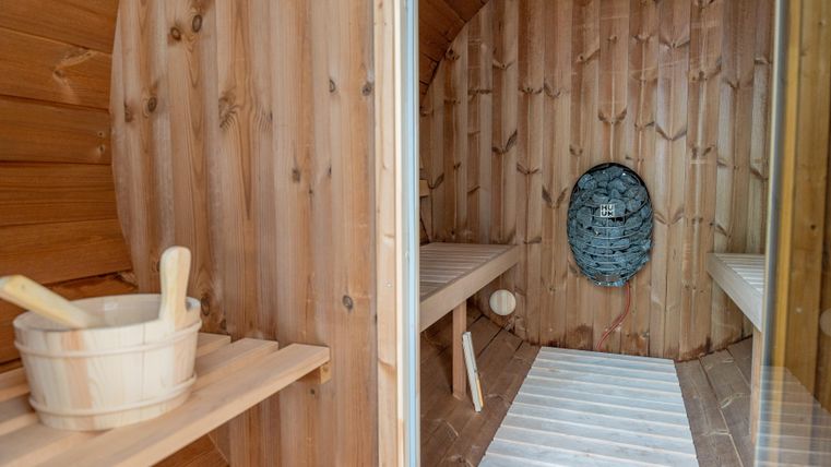 Ein gemütlicher Saunabereich aus Holz mit einer massiven Wand aus Natursteinen. Auf einer Ablage stehen ein Holzfass und eine Saunabürste.