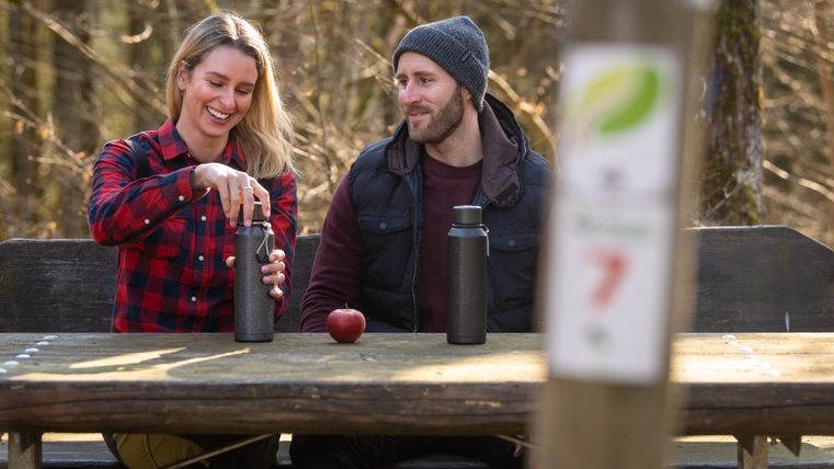 Two people are sitting at a wooden table in the forest with thermos flasks and an apple.