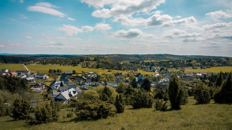 Une vue pittoresque sur un petit village avec des prairies vertes et des collines. Le ciel est clair et bleu, avec quelques nuages.